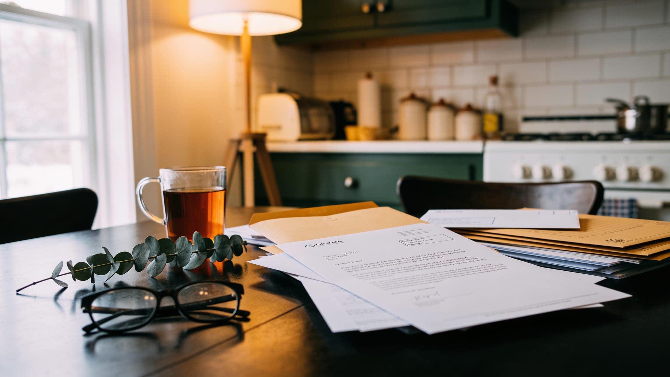 A kitchen table covered with opened envelopes, letters, reading glasses and a cup of tea.